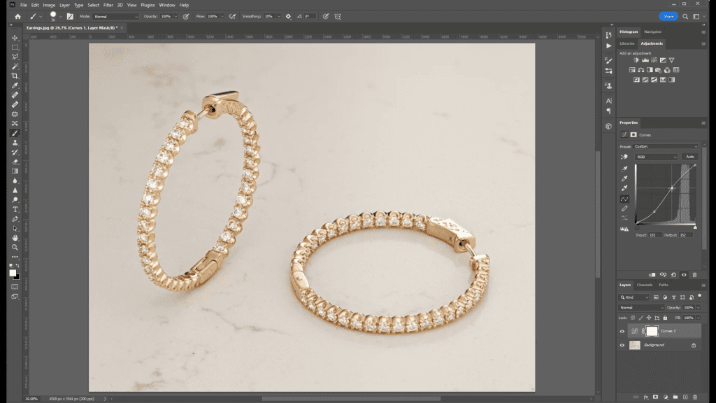 A pair of gold hoop earrings photographed in the course, lying at an angle on a light stone surface, open in Photoshop during the Adjustment Layers lesson with the Layers panel showing adjustment layers.