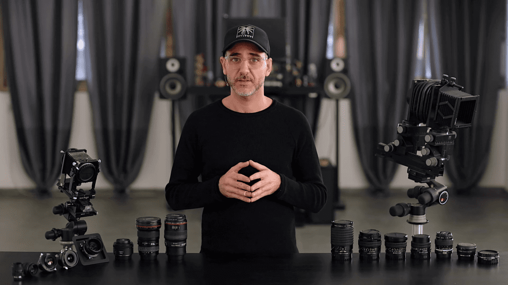 Ivan Moshe in his studio with a row of lenses and two technical view cameras on the table in front of him, from the first lesson of the Lenses chapter of Mastering Jewelry Photography.