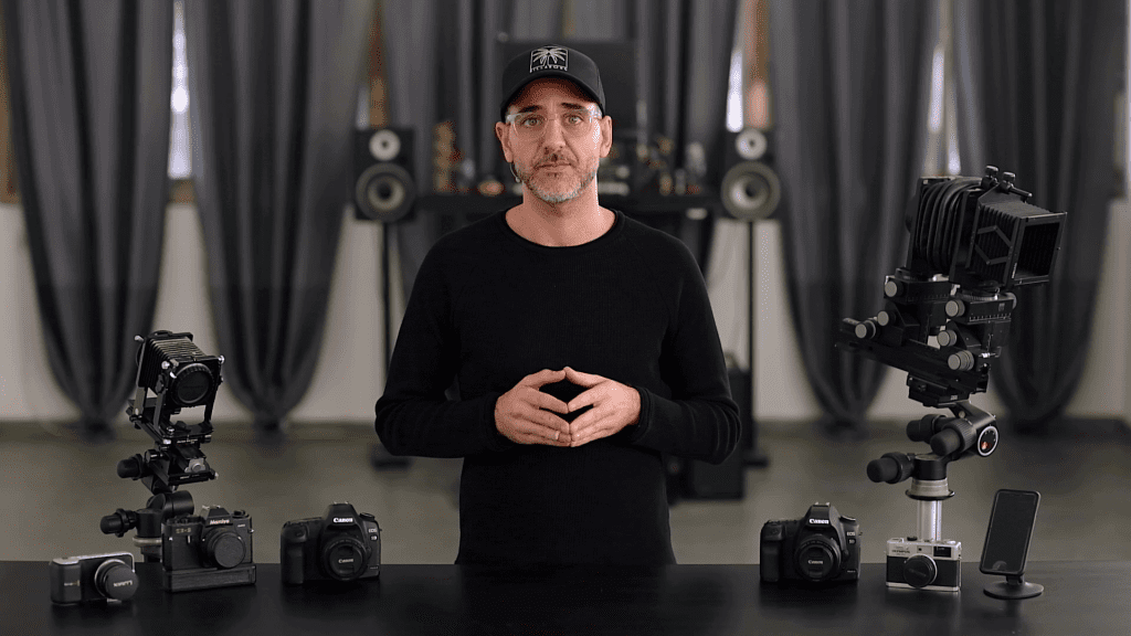 Ivan Moshe in his studio with several cameras on the table in front of him, from the first lesson of The Camera chapter of Mastering Jewelry Photography.
